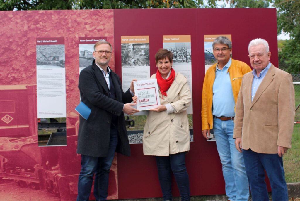 Landrat Jens Womelsdorf (li.) übergibt das Schild „Wir sind Standort der Route“ an Dr. Bettina Nickel. Über die Eröffnung des neuen Standorts der Route der Arbeits- und Industriekultur freuen sich auch der langjährige Technische Leiter des Basaltwerks, Hans Gotthard Lorch (re.), und Bürgermeister Andreas Schulz. (Foto: Landkreis Marburg-Biedenkopf/Heike Döhn)